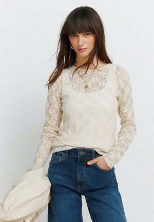 Jonge vrouw die een crème-gekleurde gehaakte top met lange mouwen draagt, gecombineerd met een hemdje, blauwe jeans, een gouden ketting en een beige jas vasthoudt.