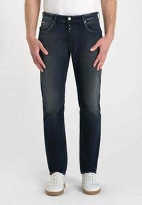 Jean denim bleu marine avec une coupe droite, présentant une légère décoloration, cinq poches et une fermeture à bouton. Porté avec des baskets blanches.