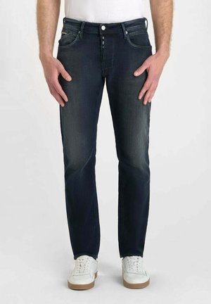 Jean denim bleu marine avec une coupe droite, présentant une légère décoloration, cinq poches et une fermeture à bouton. Porté avec des baskets blanches.