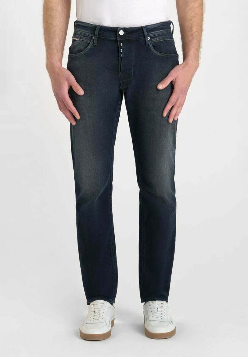 Jean denim bleu marine avec une coupe droite, présentant une légère décoloration, cinq poches et une fermeture à bouton. Porté avec des baskets blanches.