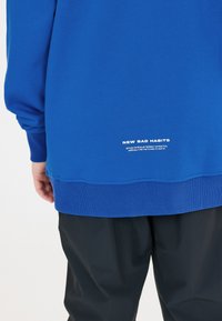 Sweatshirt bleu avec des poignets et un ourlet côtelés, arborant le texte blanc "NOUVEAUX MAUX" et un texte supplémentaire au bas de l'ourlet. Associé à un pantalon noir.