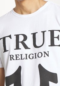 Vit bomulls t-shirt med fet svart typografi som står "TRUE RELIGION 11", med rund halsringning och korta ärmar.