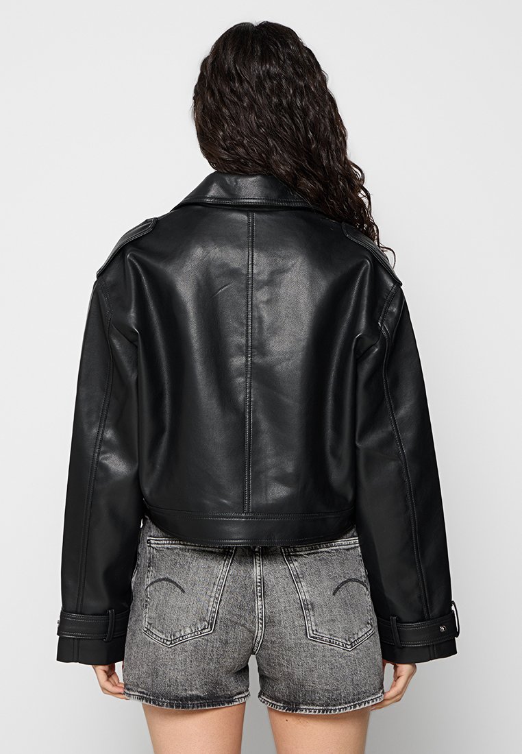 Frau mit langen lockigen Haaren, die eine schwarze Lederjacke und hochgeschnittene graue Jeansshorts trägt, steht mit dem Rücken zu einem schlichten Hintergrund.