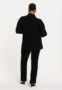 Schwarze Bluse mit Puffärmeln, geknöpftem Kragen und plissierten Details. Kombiniert mit schwarzen, figurschmeichelnden Hosen und offenen Absatzschuhen.