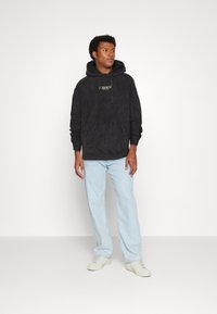 Schwarzer Hoodie mit Kängurutasche und Kordelzug, der den Schriftzug "Zodiac" in Gold trägt; kombiniert mit hellblauen geraden Jeans und weißen Sneakers.
