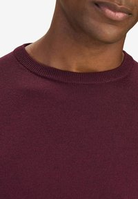 Burgundy tröja med ribbad halsringning, tillverkad av mjukt tyg. Närbild betonar texturen och den släta ytan.