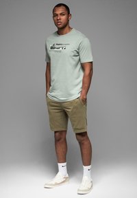 Lys grøn kortærmet t-shirt med tekst, khaki shorts med sidelagsdesign, hvide sokker og beige sneakers, stående foran en grå baggrund.