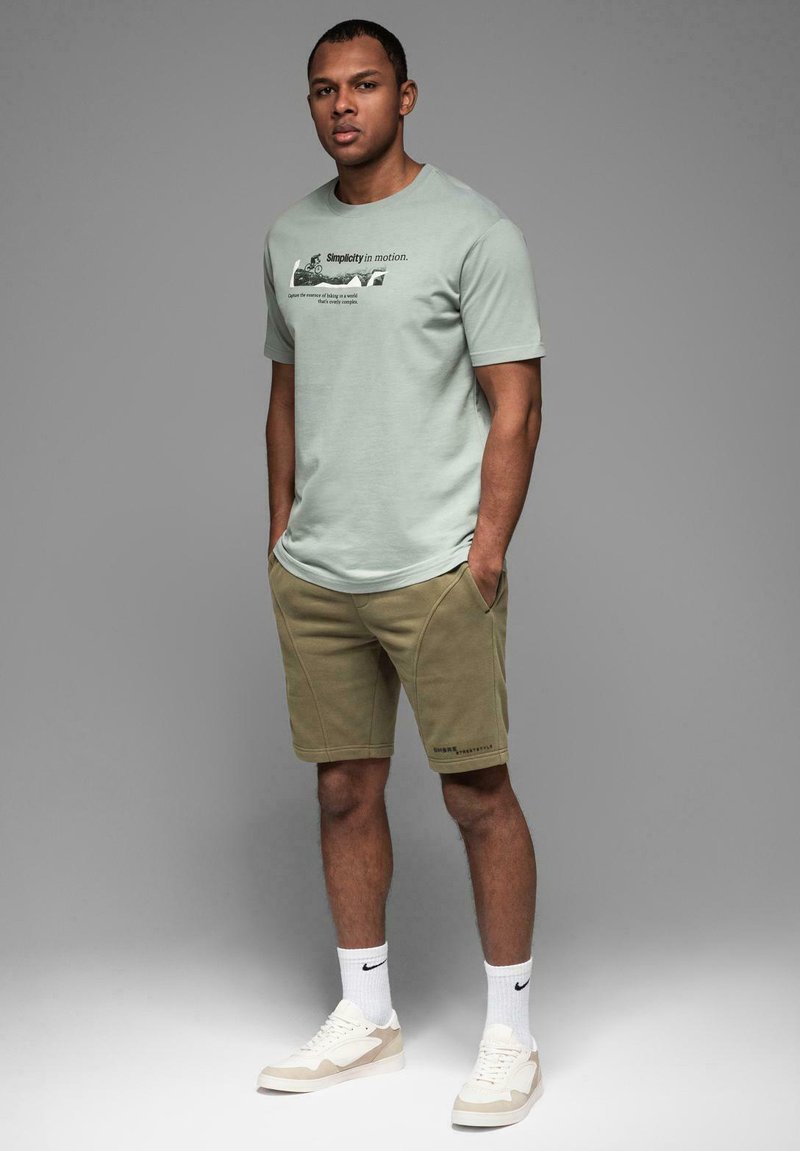 Lys grøn kortærmet t-shirt med tekst, khaki shorts med sidelagsdesign, hvide sokker og beige sneakers, stående foran en grå baggrund.