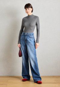 Top gris de manga larga, jeans de tiro alto y pierna ancha en azul, y zapatos rojos; sostiene un pequeño bolso burdeos. Texturas suaves, estilo casual.