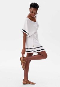 Witte jurk met zwarte zigzagafwerking, korte mouwen en een cinched taille. Tan sandalen met meerdere banden en gespsluiting. Gladde textuur.