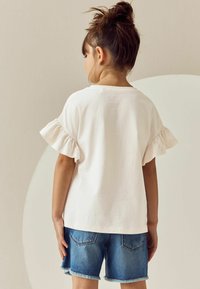 Weißes Baumwoll-T-Shirt mit gerafften kurzen Ärmeln, kombiniert mit blauen Jeans-Shorts mit ausgefranstem Saum und Gesäßtaschen.