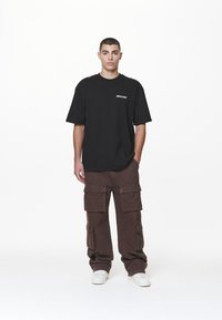 Black oversized t-shirt with logo on chest, paired with brown cargo pants featuring multiple pockets and a textured fabric. White sneakers.