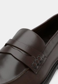 Mocassin en cuir marron à texture lisse, avec une sangle décorative et des accents métalliques. Bout arrondi et couture minimale.