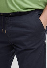 Marineblauwe katoenen broek met een tailleband met trekkoord, zijzakken en een gladde textuur, getoond met een gedeeltelijk zichtbare groene top.