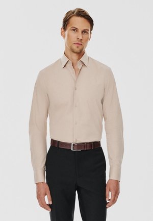Beżowa koszula z długim rękawem i guzikiem, z szpiczastym kołnierzykiem, gładką teksturą i slim fit, noszona z czarnymi spodniami i brązowym paskiem.