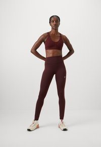 Burgunderrød sports-bh med teksturert mønster, parret med matchende høy midje leggings. Modell iført beige treningssko med svarte detaljer.