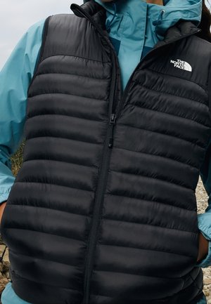 Person, die eine schwarze gesteppte North Face-Weste über einer blauen Jacke trägt und an einem bewölkten Tag im Freien steht.