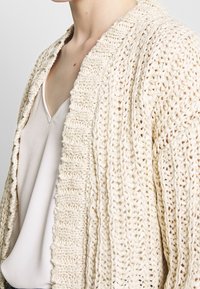 Gebreide cardigan in gebroken wit, met een chunky textuur en brede open voorkant. Gedragen over een lichtgrijze v-hals top met dunne bandjes.