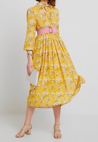 Robe midi florale jaune avec de longues manches en cloche, ceinture rose cintrée et jupe fluide. Les accessoires incluent une pochette blanche et des sandales nudes.