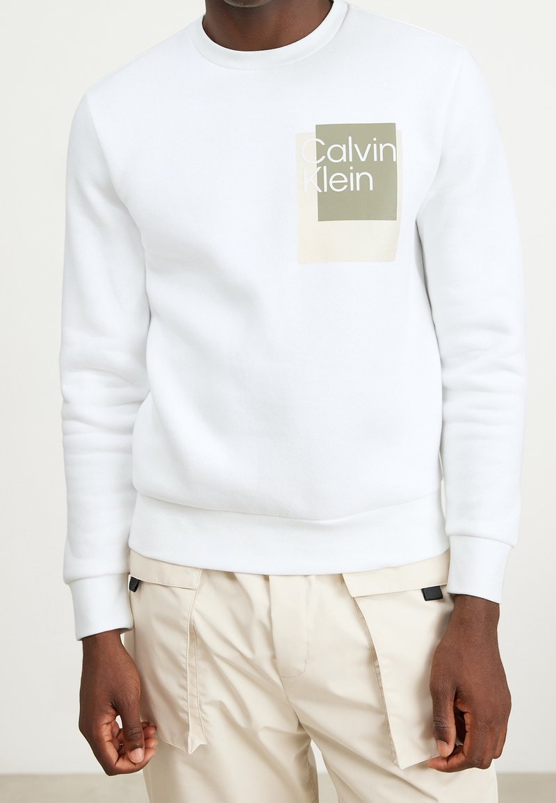 Sudadera blanca con puños y dobladillo acanalados. En la parte delantera presenta un gráfico rectangular con "Calvin Klein" en blanco sobre un fondo verde y beige.