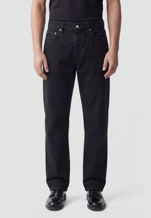Persona che indossa jeans neri a gamba dritta e scarpe in pelle nere, in piedi davanti a uno sfondo chiaro e semplice.