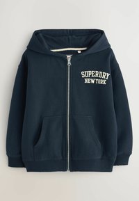 Sudadera navy con cremallera y capucha con cordón, bolsillos frontales, y el logo "SUPERDRY NEW YORK" en crema en el lado izquierdo del pecho. Textura suave, puños acanalados.