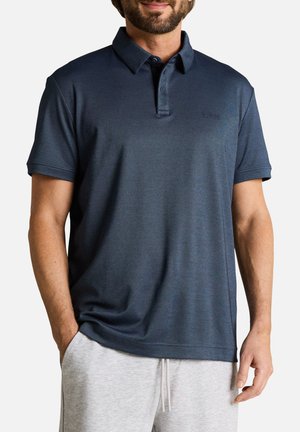 Homme portant un polo bleu marine à manches courtes et un pantalon gris clair à cordon, avec une main dans la poche.