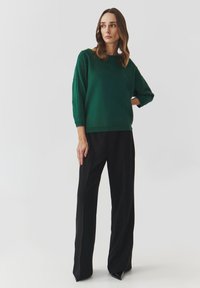 Maglione verde con maniche a tre quarti, dettagli testurizzati e scollo a girocollo abbinato a pantaloni larghi neri. Finitura del tessuto liscia.