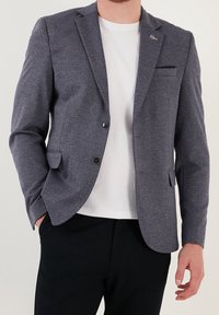 Mężczyzna w szarym żakiecie z dwoma guzikami, pod którym ma białą koszulę i czarne spodnie, stoi z jedną ręką w kieszeni.