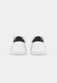 Een paar witte sneakers met zwarte hieltabs, met 'Calvin Klein Jeans' embossed op de witte rubberen zolen, gezien van de achterkant tegen een witte achtergrond.