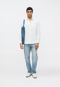 Camisa de linho branca com botões e bolso, jeans azul claro, e uma bolsa tote de ganga azul, usados com sapatilhas brancas sobre um fundo simples.