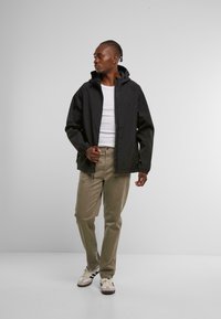 Chaqueta ligera negra con capucha, cierre de cremallera y bolsillos laterales. Llevada sobre una camisa blanca, combinada con pantalones verde claro y zapatillas.