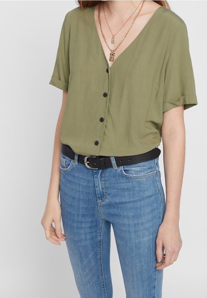 Femme portant un chemisier vert olive à manches courtes boutonné, rentré dans un jean bleu avec une ceinture noire, et des colliers dorés superposés.
