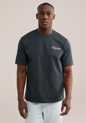 Muž v tmavosivej tričku s nápisom "Downtown Social Club" a svetlomodrých džínsoch stojí pred jednofarebným neutrálnym pozadím.