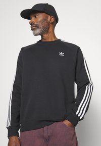 Crna dukserica s bijelim dizajnom trostrikaste linije duž rukava, rebrastim manžetama, okruglim izrezom i malim Adidas logotipom na prsima.