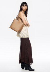 Brązowa torebka typu tote ze skóry, z podwójnymi uchwytami, gładką fakturą i subtelnym, wytłaczanym logo. Modelka ma na sobie białą bluzkę i brązową spódnicę wykończoną koronką oraz czarne buty.