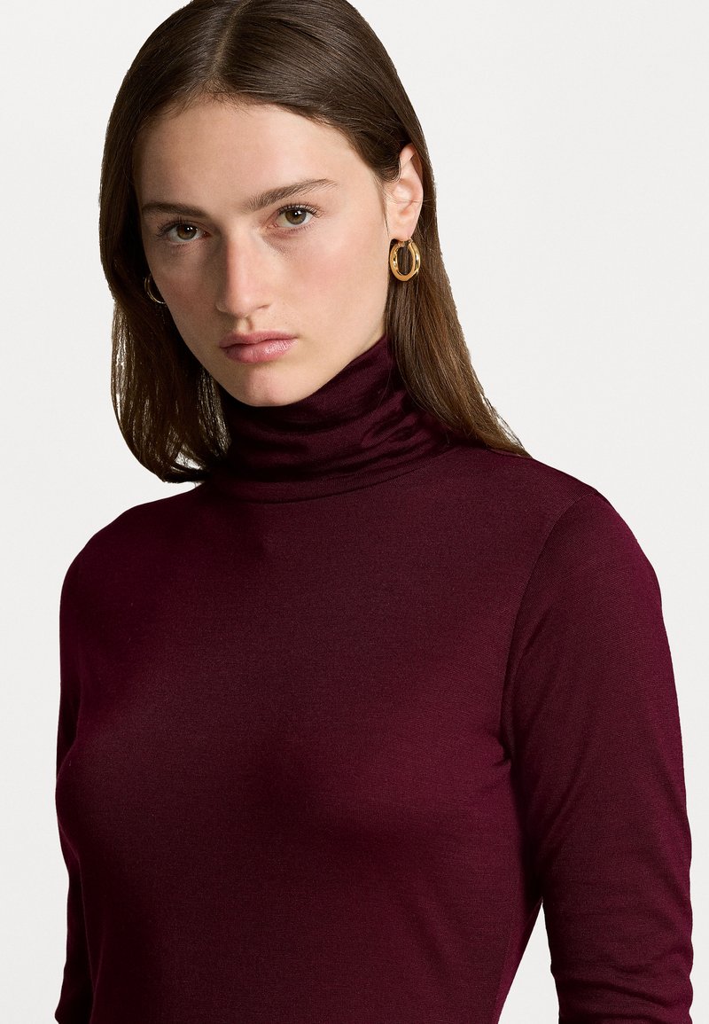 Burgundrot Turtleneck-Oberteil aus weichem Stoff, taillierter Schnitt, mit hohem Kragen und langen Ärmeln, ergänzt durch goldene Creolen.