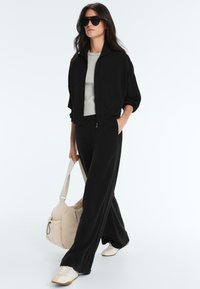 Femme portant une veste noire, un pantalon noir à jambes larges, des baskets blanches, des lunettes de soleil, portant un sac beige, marchant sur un fond blanc uni.