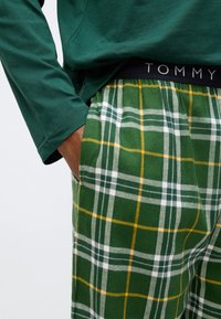 Πράσινο μακρυμάνικο πουκάμισο συνδυασμένο με πράσινες καρό φόρμες jogger που έχουν κίτρινες και λευκές γραμμές, με ελαστική μέση που φέρει την ετικέτα "TOMMY."