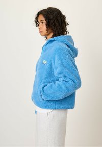Blauwe fleece hoodie met een cropped ontwerp, voorzien van een capuchon en een rits aan de voorkant. Zachte textuur en twee zijzakken, met een logo patch.