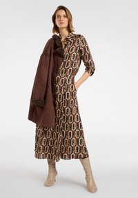 Largo vestido marrón con estampado y mangas hasta el codo, con detalle en la cintura, combinado con un abrigo de ante marrón con acentos de piel. Botas beis.