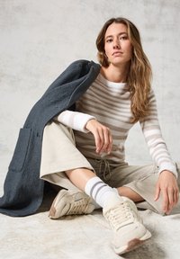 Maglione a righe beige e bianco, pantaloni beige a gamba larga, cappotto grigio appoggiato, calzini bianchi con righe marroni e sneakers beige.