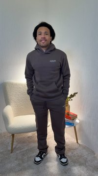 Σκούρα γκρι φούτερ με κουκούλα και παντελόνι φόρμας από απαλό ύφασμα, με μπροστινή τσέπη και διακριτικό λογότυπο. Μαύρα και λευκά αθλητικά παπούτσια ολοκληρώνουν το σύνολο.