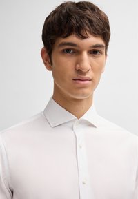 Chemise blanche à manches longues avec un col classique, texture lisse et petits boutons. Le tissu semble léger et légèrement brillant.