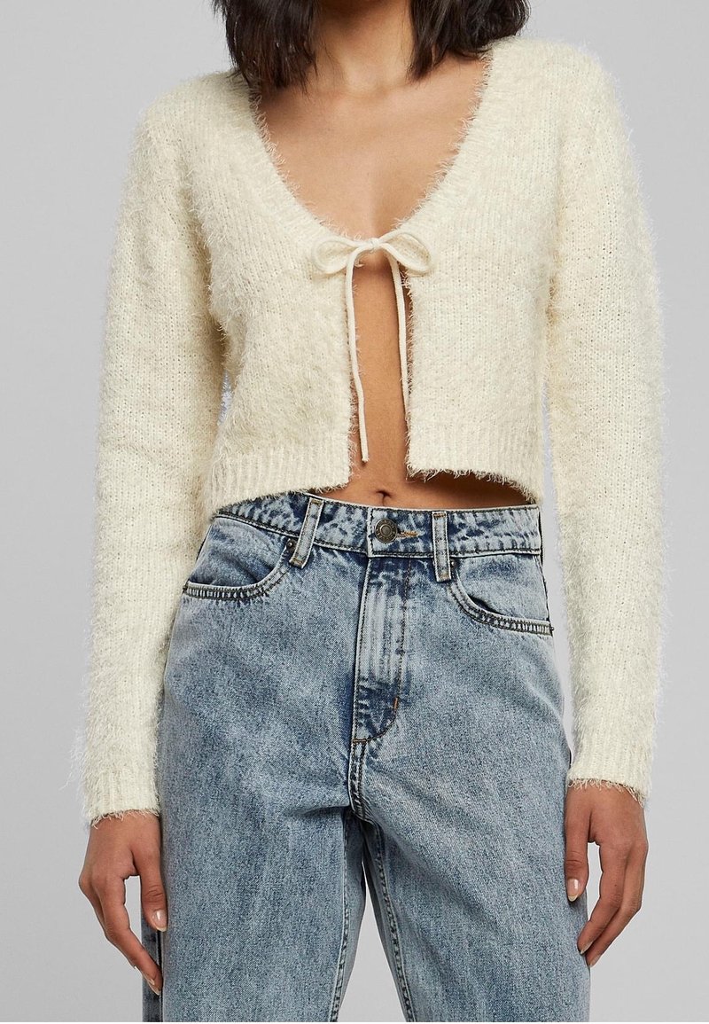 Cream, fuzzy cropped sweater with a deep V-neck and tie closure at the front, paired with light blue high-waisted denim jeans.
