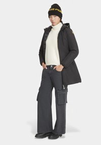 Giacca a piumino nera con cappuccio, dolcevita crema, pantaloni cargo grigi a gamba larga con tasche e stivali neri, abbinati a un berretto nero.