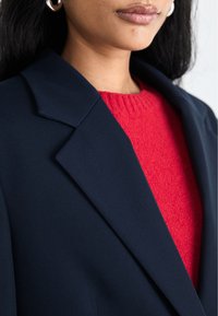 Blazer marine à revers pointus assorti à un pull en tricot texturé rouge. Mettez en avant le contraste entre le blazer lisse et le haut douillet.
