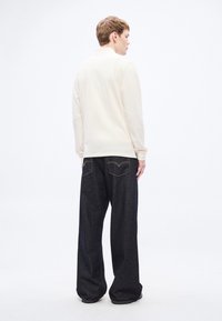 Gaiši bēša garām piedurknēm krekls ar apkakli, kombinēts ar tumšām platām džinsa džinsiem ar kontrastējošām šuvēm un aizmugurējām kabatām.