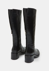 Botas negras hasta la rodilla con un acabado liso y brillante. Cuentan con una cremallera lateral, un tacón grueso y un detalle de costura mínima.