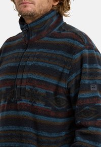 Fleece pullover met hoge kraag, voorzien van donkerblauwe, bruine en teal strepen. Inclusief een ritszak op de borst en een logopatch op de mouw.